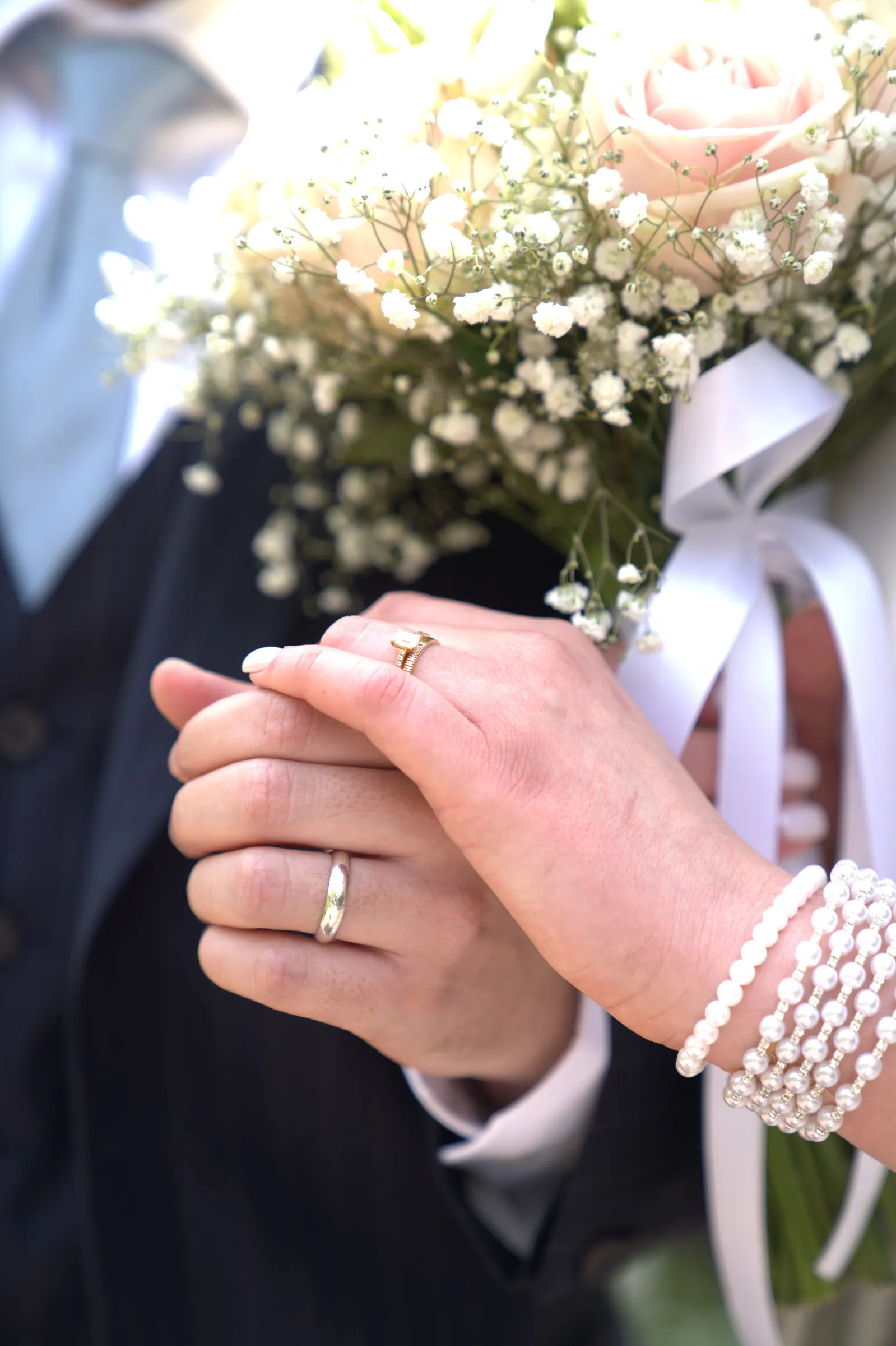 Les mains des mariés réunies devant le bouquet, alliances et bracelet de perles
