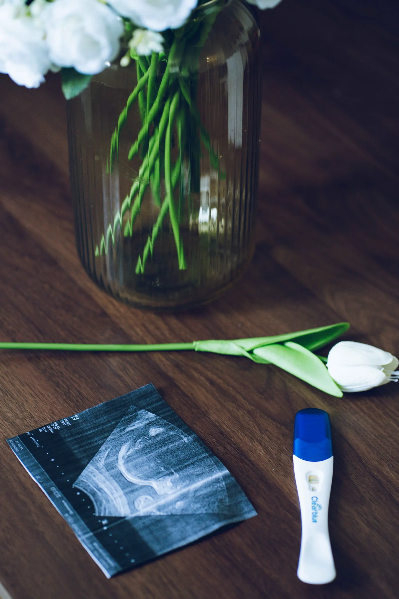 Un test de grossesse et des échographies posés sur une table, à côté d’un vase de fleurs