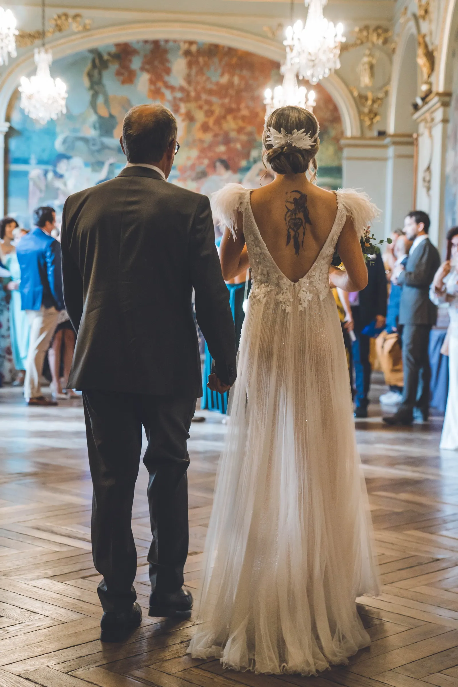 Un père accompagne la mariée dans la salle, vus de dos, robe au dos nu