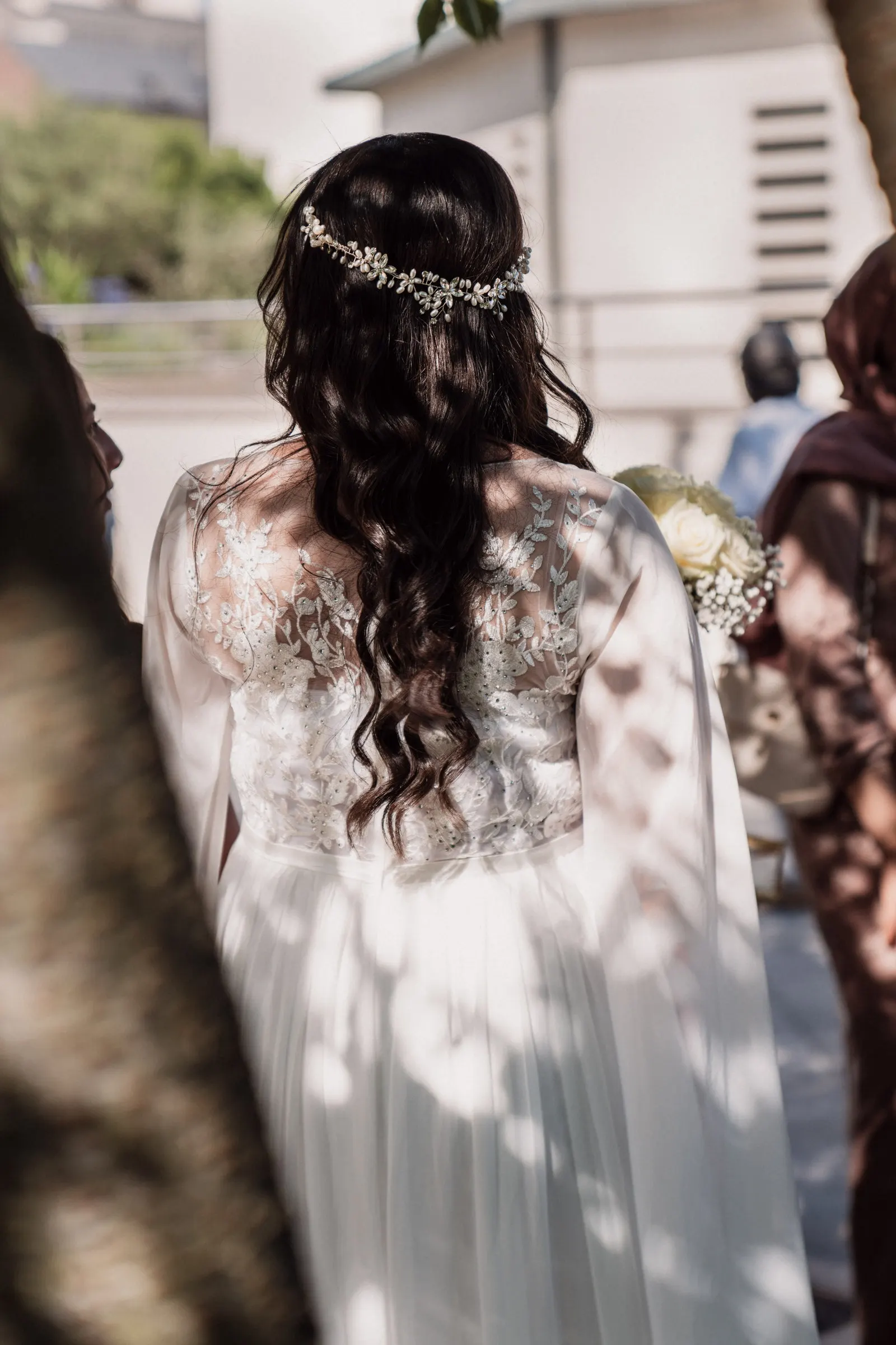 La mariée de dos, un bijou de tête fleuri dans les cheveux, la lumière filtrée par les feuilles
