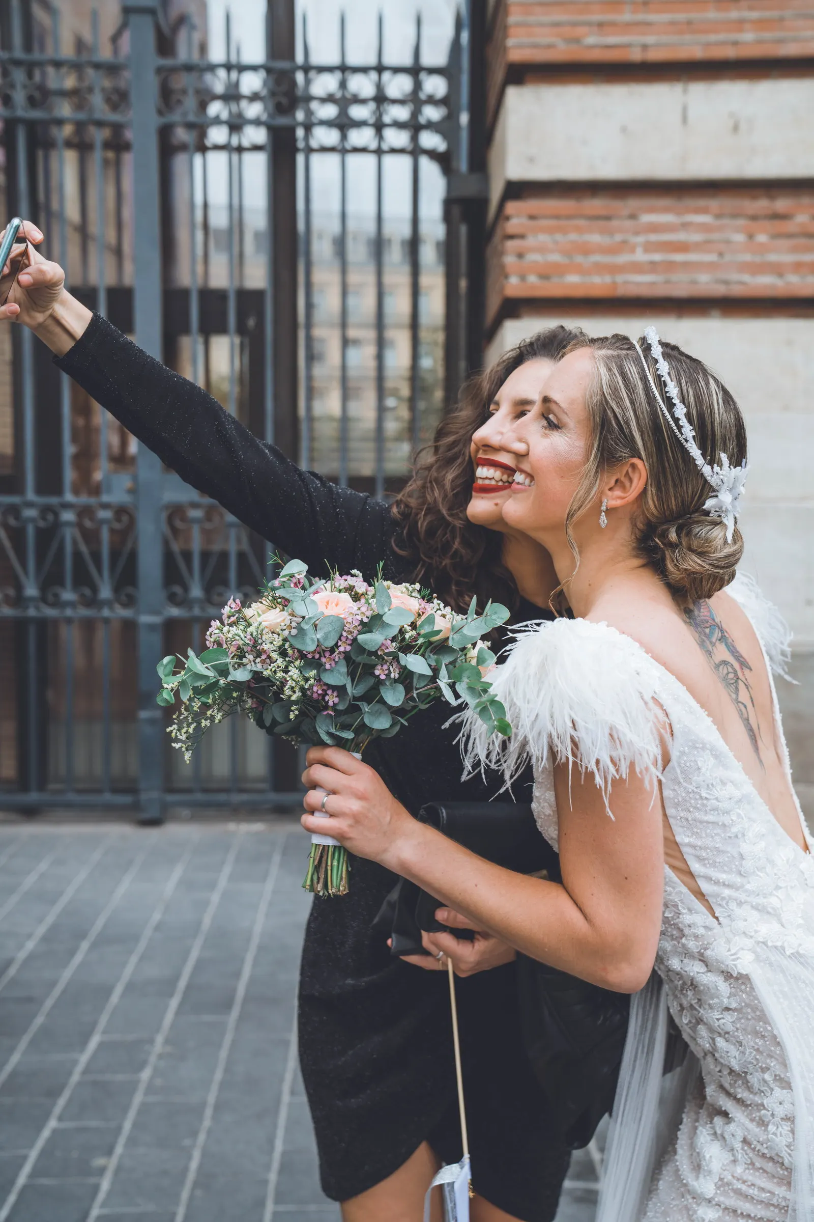 La mariée rit, un bras levé, son bouquet à la main, devant une grille en fer